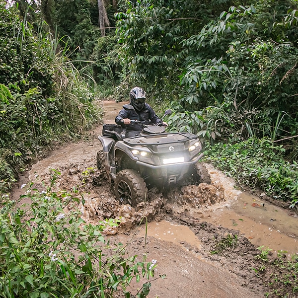 Piloto em quadriciclo Can-Am passando pela poça de lama em uma trilha de terra.